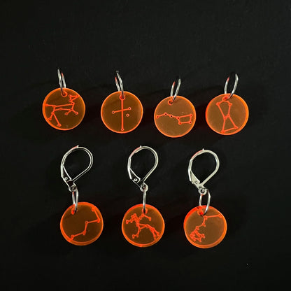 Seven orange circular perspex stitch markers with various constellation designs, four attached to leverback hooks and three on jump rings, arranged on a black background.