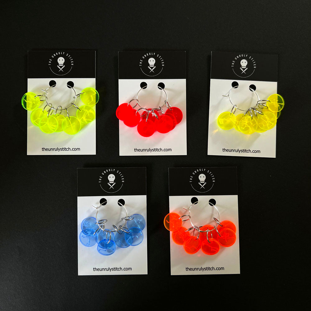 Five sets of perspex stitch markers packaged on cards from "The Unruly Stitch," each set containing multiple stitch markers in neon yellow, red, blue, and orange.