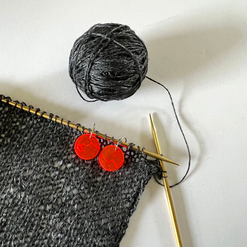 Close-up of a black knitted fabric on a gold knitting needle, with a ball of gray yarn beside it. Two red circular perspex stitch markers with constellation designs sit on the knitting needle.