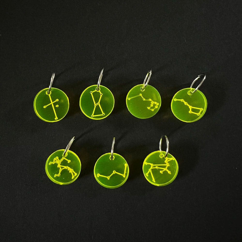 Seven neon yellow circular perspex stitch markers with various constellation designs on silver jump rings, arranged on a black background.