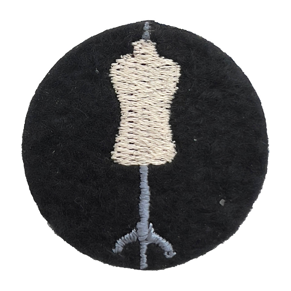 Close-up of a felt badge with an embroidered dressmaker's mannequin in beige and gray threads on a black background, designed by The Unruly Stitch.