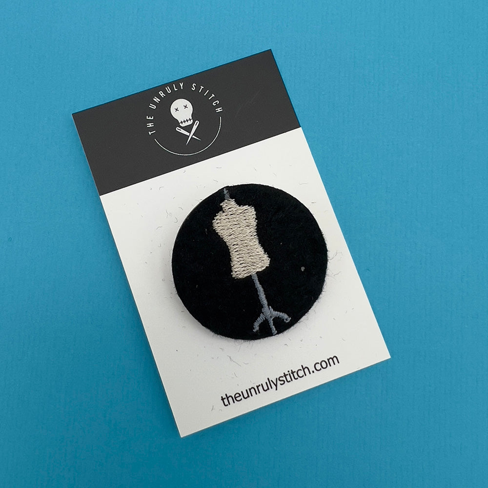 Embroidered felt badge with a dressmaker's mannequin design in beige and gray threads on a black background, mounted on a branded card from The Unruly Stitch.