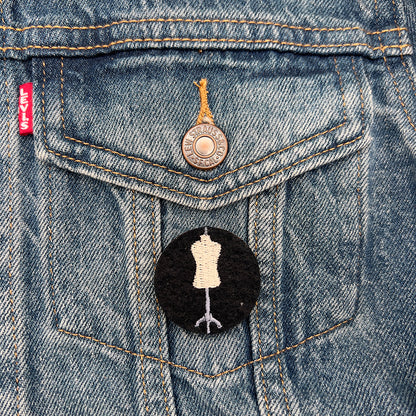 Embroidered felt badge depicting a dressmaker's mannequin in beige and gray threads on a black background, pinned on the pocket of a denim jacket, designed by The Unruly Stitch.