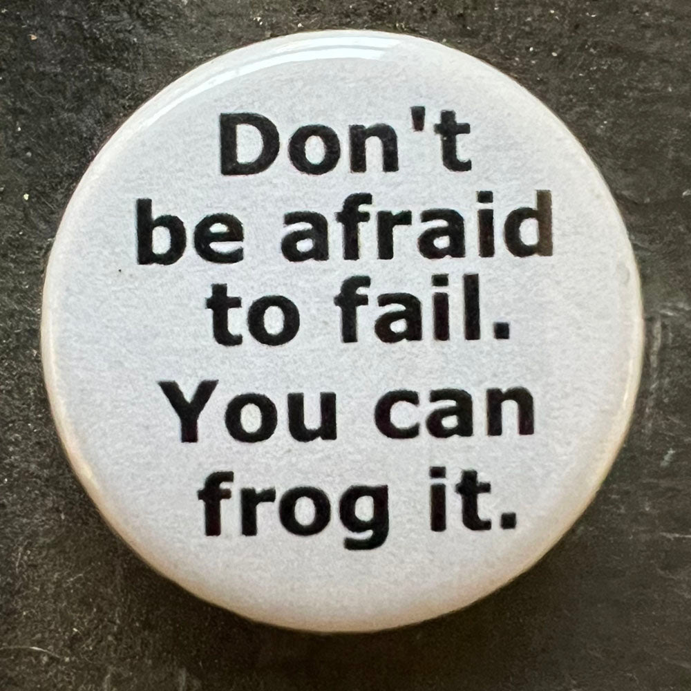 Close-up of a white Don't be afraid to fail. You can always frog it pin badge with black text.