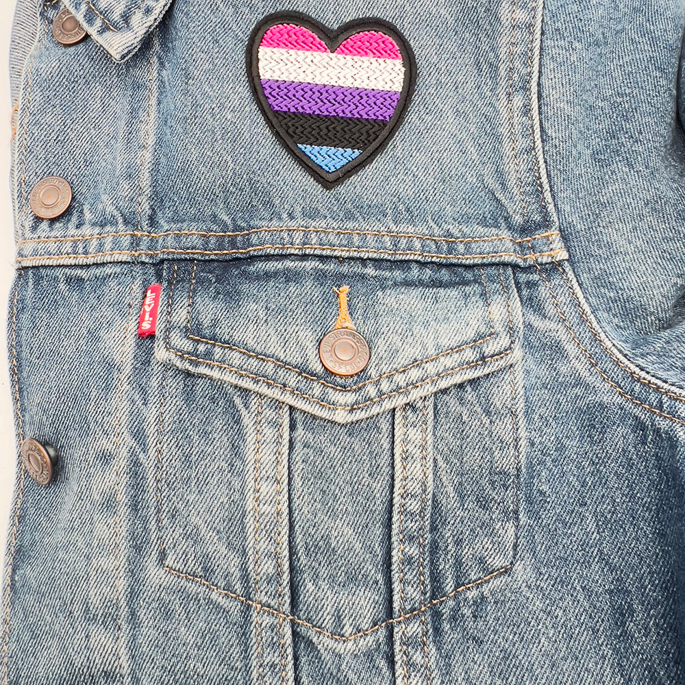 Gender fluid pride flag heart-shaped embroidered felt patch attached to the front pocket of a denim jacket. The patch displays horizontal stripes in pink, white, purple, black, and blue.