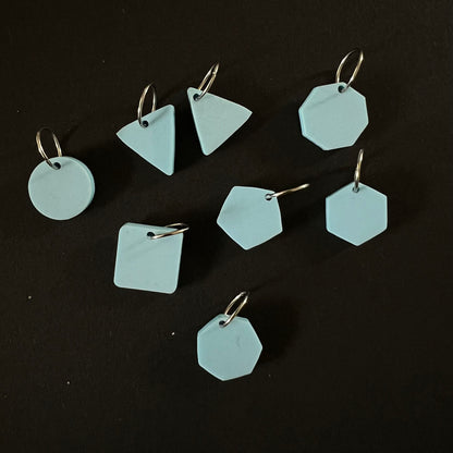 A set of geometric stitch markers in light blue perspex on jump rings, including circles, triangles, squares, pentagons, hexagons, and octagons, displayed on a black background.