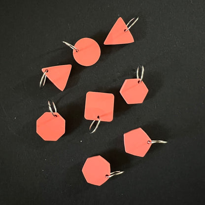 A set of geometric stitch markers in coral perspex on jump rings, including circles, triangles, squares, pentagons, hexagons, and octagons, displayed on a black background.