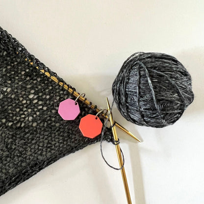 A close-up of a knitting project in progress using dark gray yarn, with two stitch markers attached to the knitting needles – one octagon-shaped in coral perspex and one in lavender perspex.