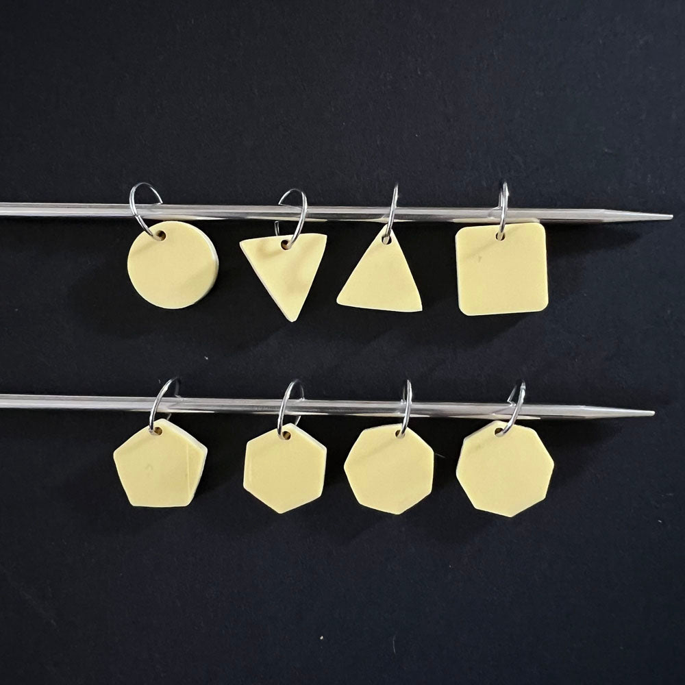 geometric stitch markers in pale yellow perspex on jump rings, displayed on a knitting needle. The shapes include circles, triangles, squares, pentagons, hexagons, and octagons, against a black background.