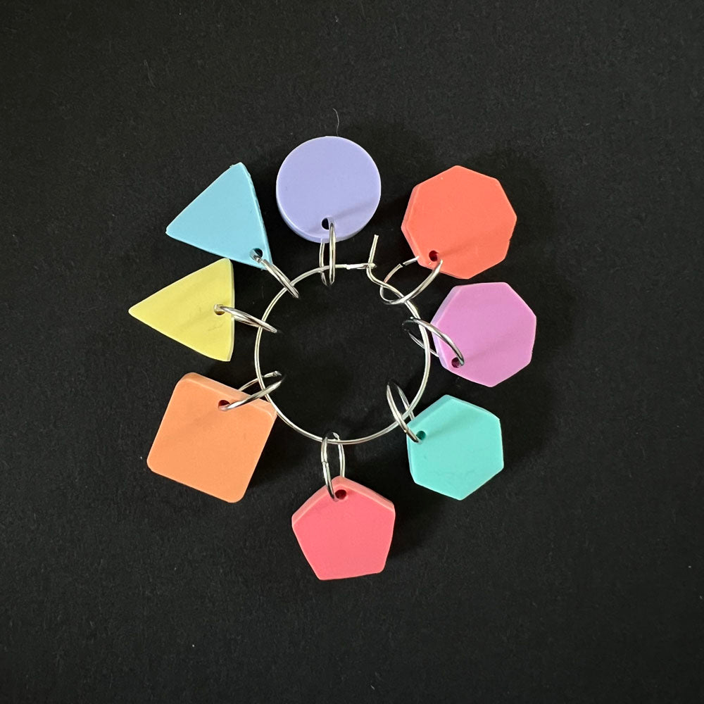 A ring of geometric stitch markers in assorted pastel colors, including yellow, orange, green, blue, purple, and pink, arranged in a circle on a black background.