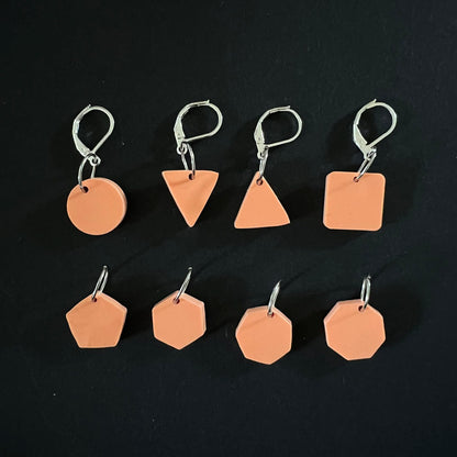 A set of geometric stitch markers in terracotta perspex on jump rings, including circles, triangles, squares, pentagons, hexagons, and octagons, displayed on a black background.