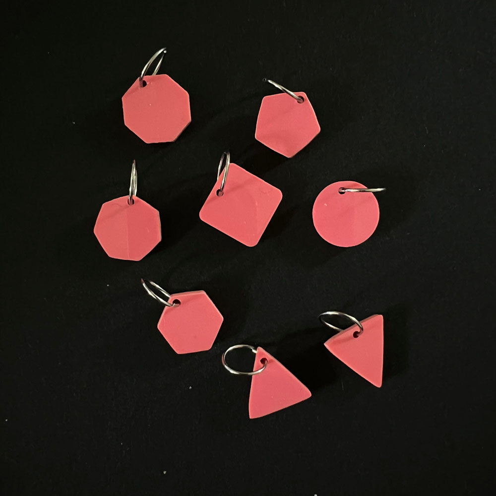 A set of geometric stitch markers in red perspex on jump rings, including circles, triangles, squares, pentagons, hexagons, and octagons, displayed on a black background.
