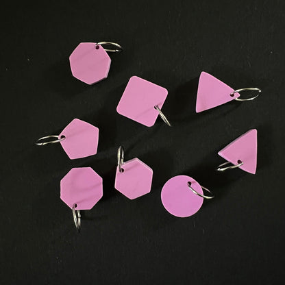 A set of geometric stitch markers in raspberry perspex on jump rings, including circles, triangles, squares, pentagons, hexagons, and octagons, displayed on a black background.