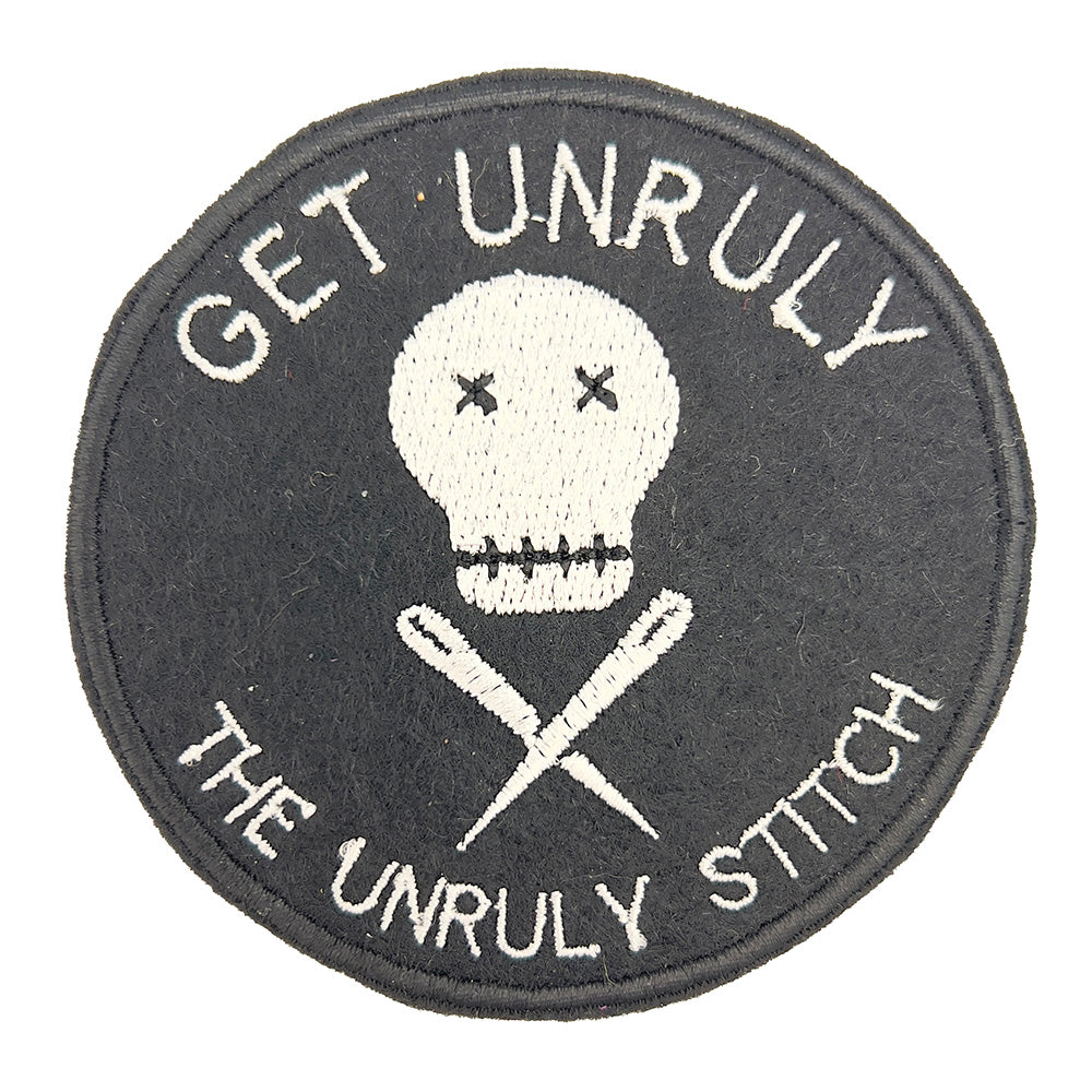 Close-up image: Close-up view of an embroidered felt patch with the text "Get Unruly The Unruly Stitch" surrounding a white skull and crossed needles. The patch is circular and features white embroidery on a black background.