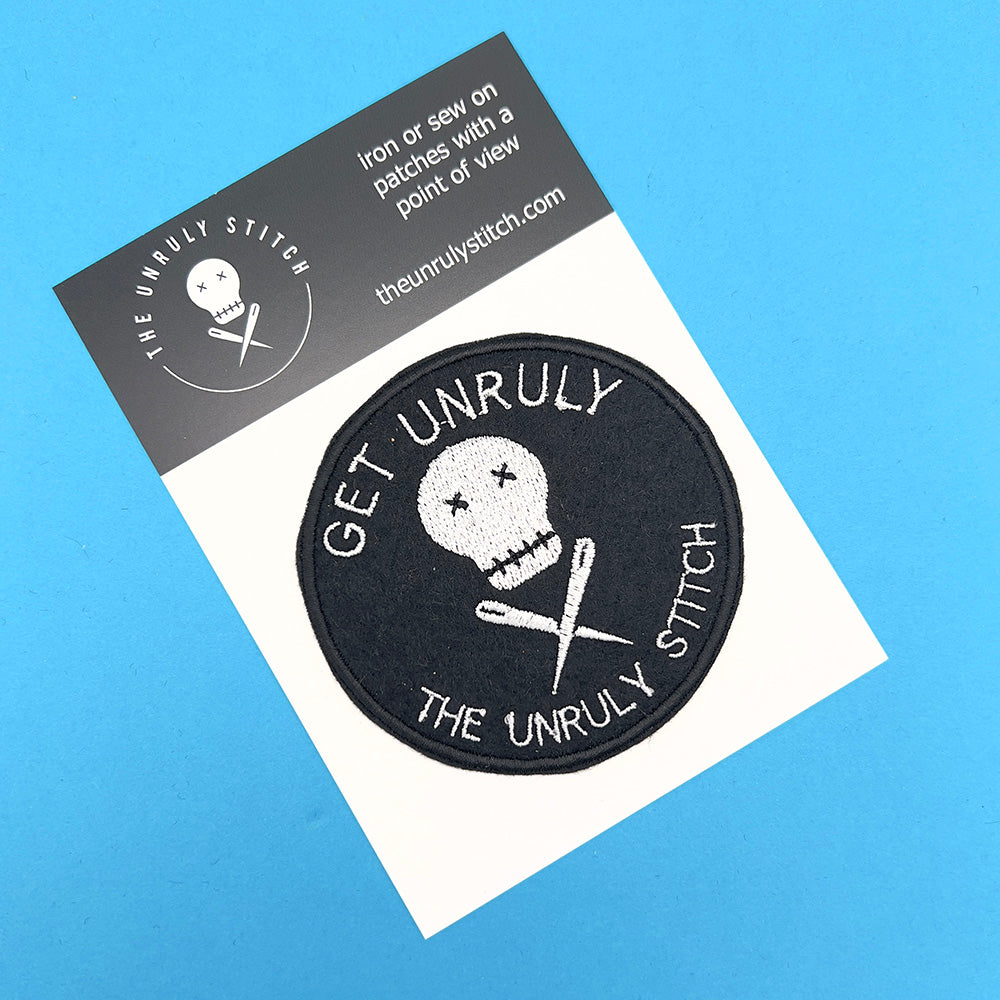 On card image: An embroidered felt patch with the text "Get Unruly The Unruly Stitch" surrounding a white skull and crossed needles is displayed on a card. The patch is circular and features white embroidery on a black background. The card has the logo and website of The Unruly Stitch.