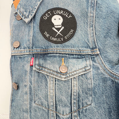 On denim jacket image: An embroidered felt patch with the text "Get Unruly The Unruly Stitch" surrounding a white skull and crossed needles is sewn onto the front pocket of a denim jacket. The patch is circular and features white embroidery on a black background.