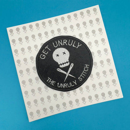 A round black patch with white embroidery featuring a skull and knitting needles, and the text 'GET UNRULY THE UNRULY STITCH'. The patch is placed on a gift card with a repeating skull and knitting needles pattern.