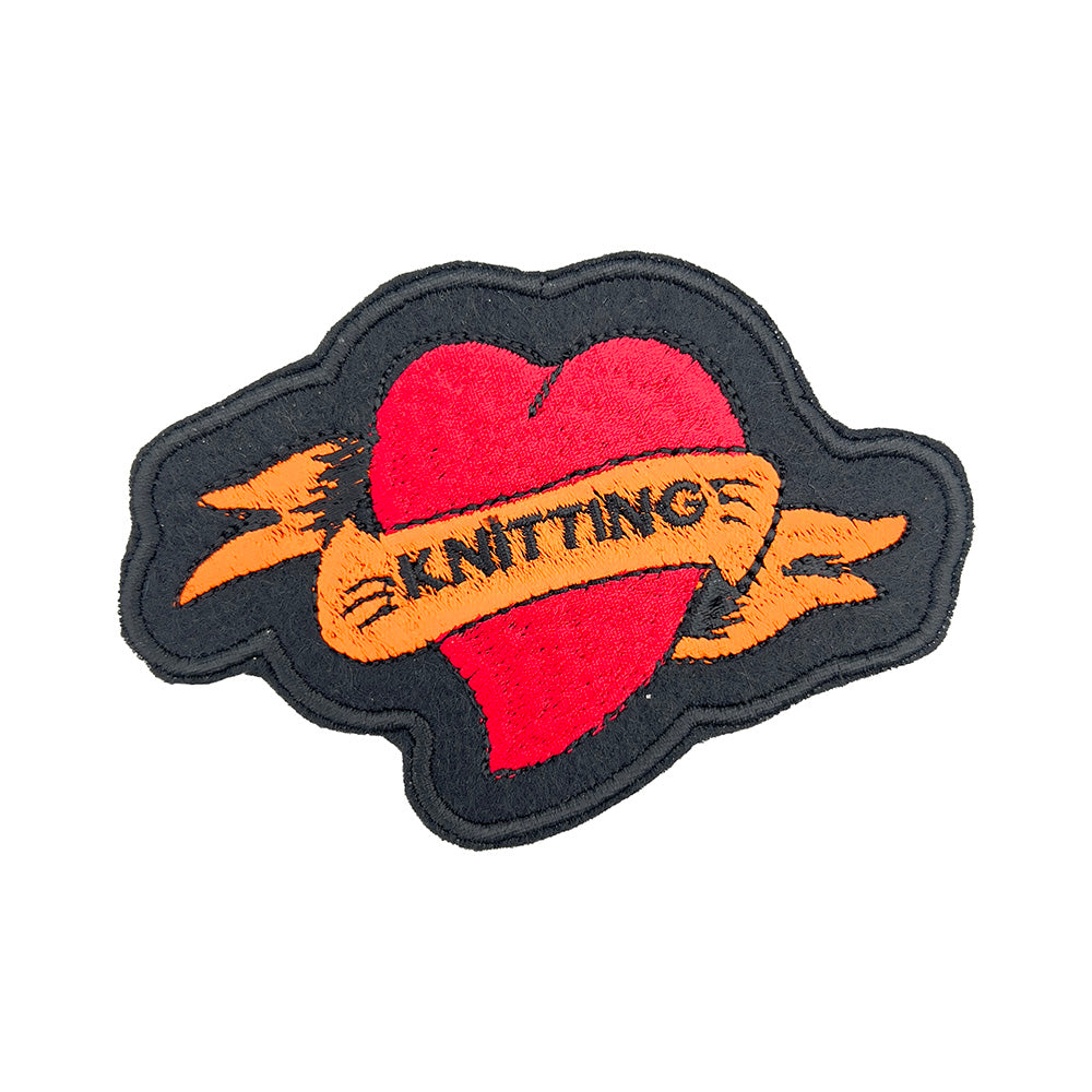 Close-up image: Close-up view of an embroidered felt patch in the shape of a red heart with an orange banner. The banner has the word "KNITTING" stitched in black, and the heart and banner are outlined in black.