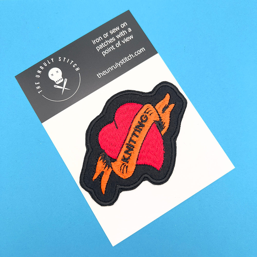 Image of an embroidered felt patch in the shape of a red heart with an orange banner on a product display card. The banner reads "KNITTING" in black stitching, and the heart and banner are outlined in black. The card has the text "The Unruly Stitch" and instructions for ironing or sewing on patches.