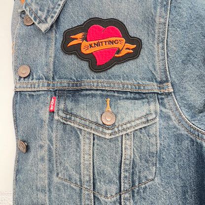 Image of an embroidered felt patch in the shape of a red heart with an orange banner attached to a denim jacket. The banner has the word "KNITTING" stitched in black, and the heart and banner are outlined in black, positioned above the jacket pocket.