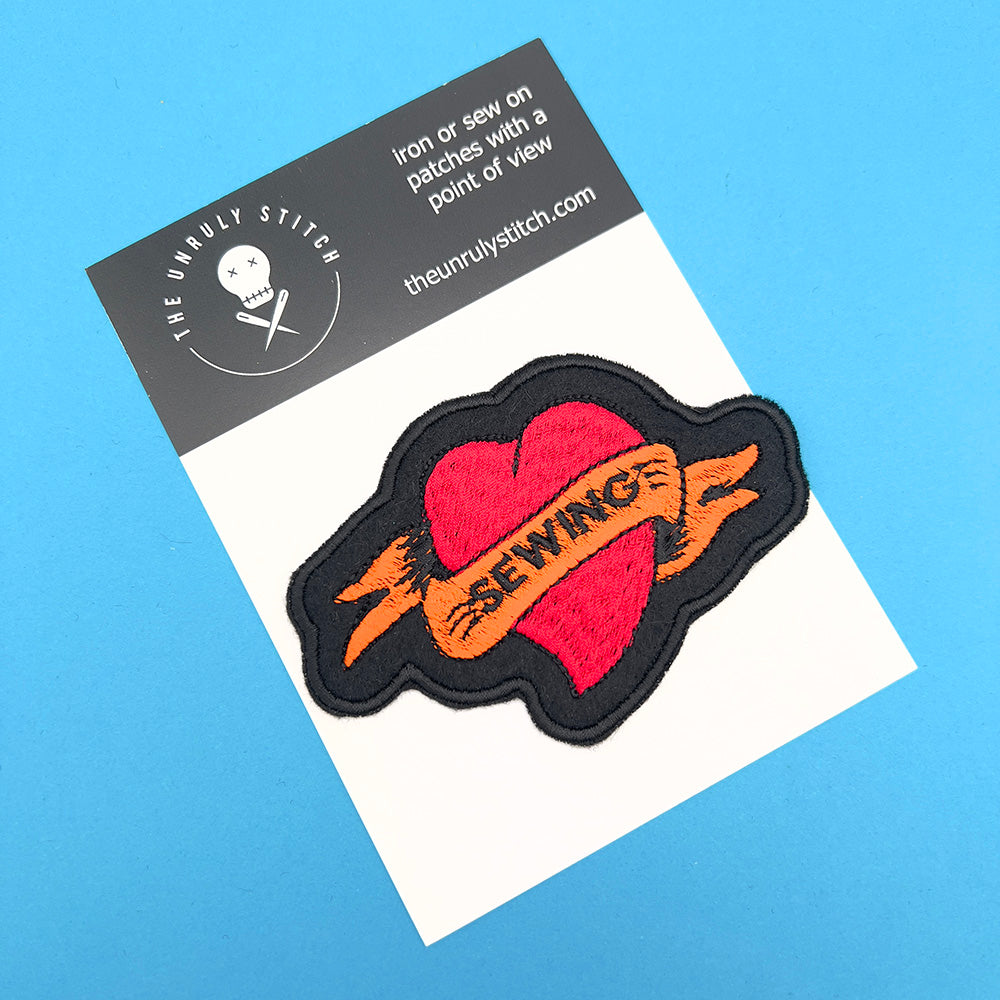 On card image: An embroidered felt patch shaped like a heart with a banner, displayed on a card. The patch features a red heart with an orange banner that reads "SEWING," with black outlines and details.