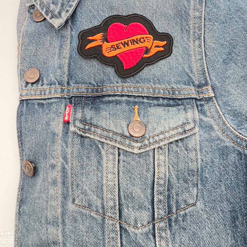 On jacket image: An embroidered felt patch shaped like a heart with a banner, attached to a denim jacket. The patch features a red heart with an orange banner that reads "SEWING," with black outlines and details.
