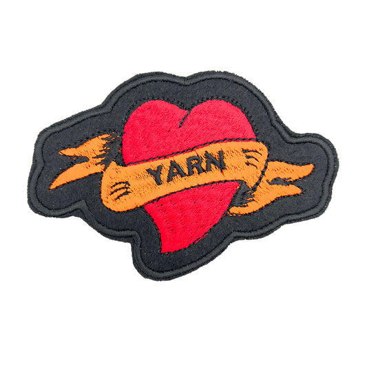 Close-up image: Close-up view of an embroidered felt patch shaped like a heart with a banner. The patch features a red heart with an orange banner that reads "YARN," with black outlines and details.