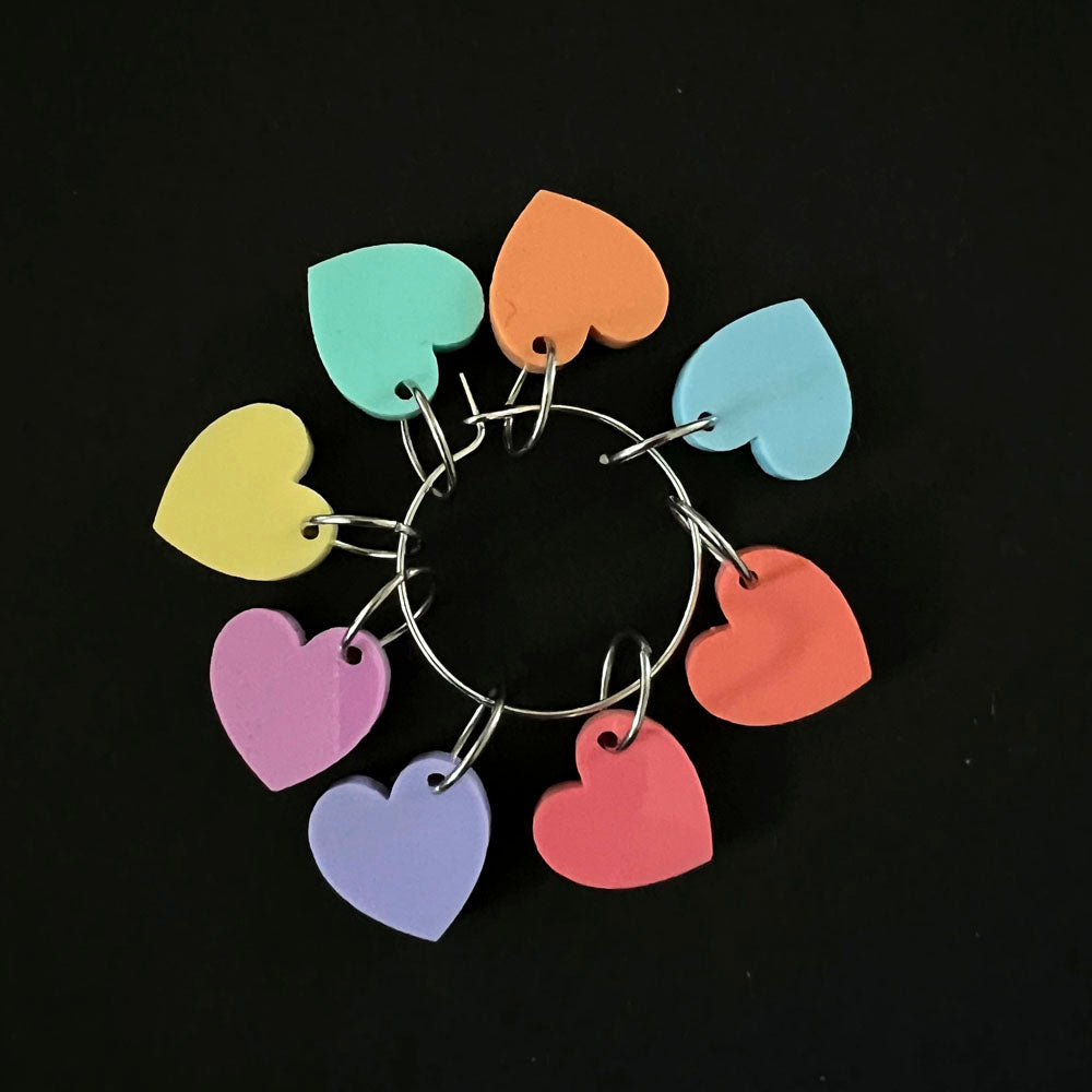 A circular arrangement of heart-shaped stitch markers in various pastel colors, including yellow, green, blue, purple, pink, orange, and red, all attached to a central metal ring.