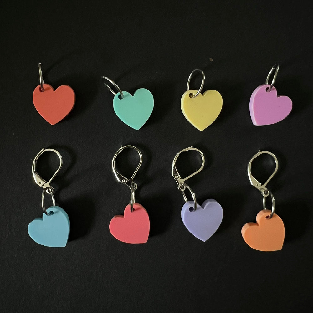 A collection of eight heart-shaped stitch markers in pastel colors, each marker displayed individually with a mix of jump rings and latch-back hooks, against a black background.