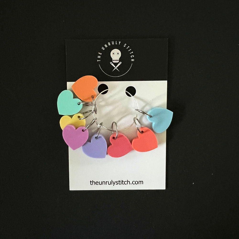 A set of heart-shaped stitch markers in pastel colors displayed on a branded card from "The Unruly Stitch," showing the markers attached to a central metal ring.