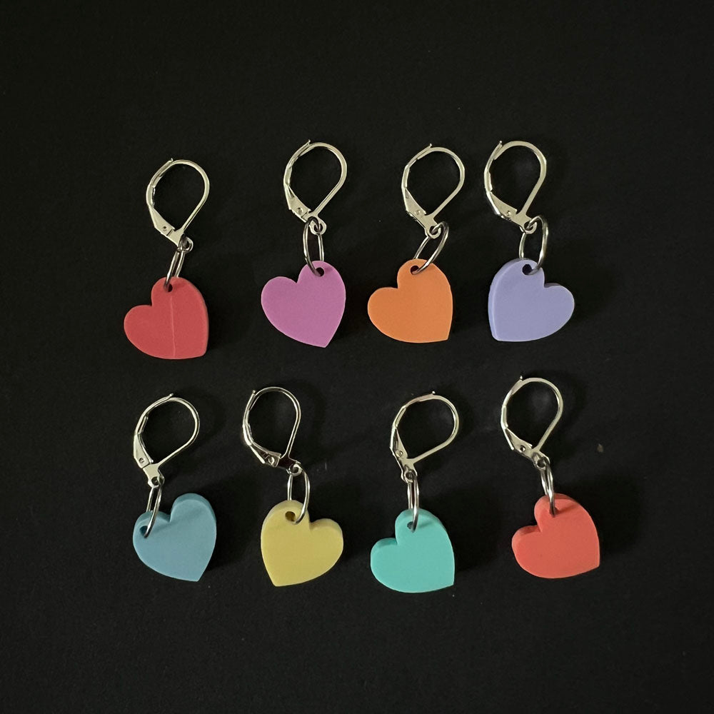 Another arrangement of eight heart-shaped stitch markers in pastel colors, each marker displayed individually with latch-back hooks, laid out in two rows against a black background.