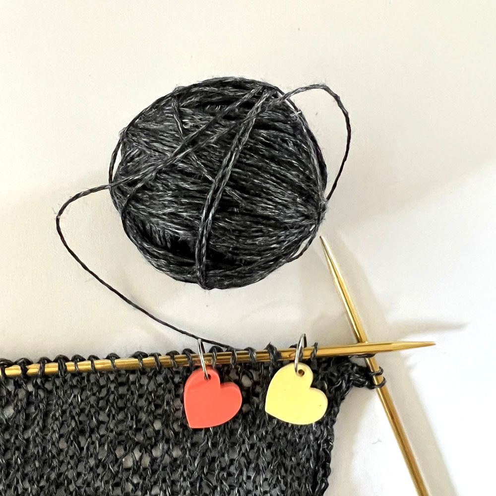 Two heart-shaped stitch markers, one red and one yellow, attached to a piece of knitting with golden knitting needles, alongside a ball of black and gray yarn.
