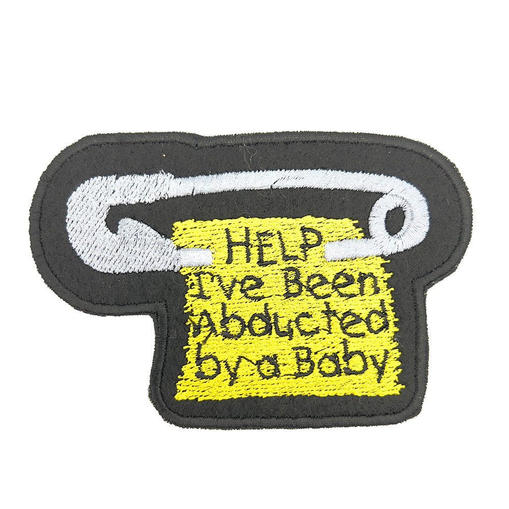 Close-up image of a felt patch shaped like a safety pin with a yellow tag hanging from it. The tag has the text "HELP I've Been Abducted by a Baby" embroidered in black.The patch is embroidered onto black felt fabric with a black satin stitch border. the-patch-is-on-a-plain-white-background 