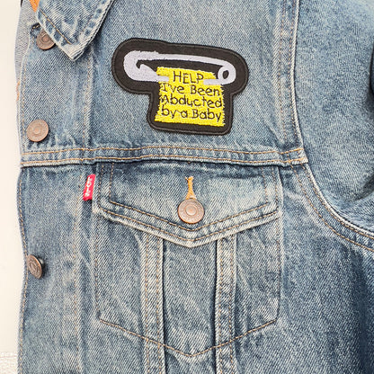 The same safety pin-shaped patch with the text "HELP I've Been Abducted by a Baby" is ironed onto a blue denim jacket, positioned above the pocket on the left side of the jacket.