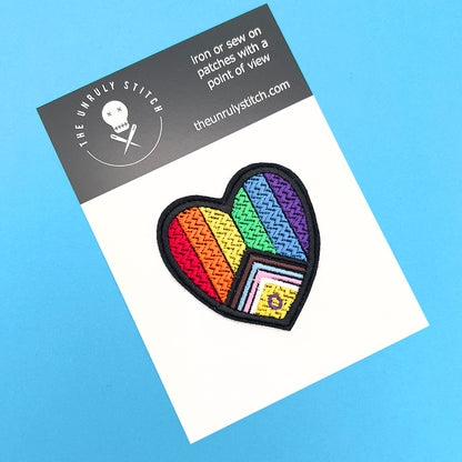 Heart-shaped Inclusive Pride flag embroidered patch displayed on a branded card from The Unruly Stitch, against a blue background.