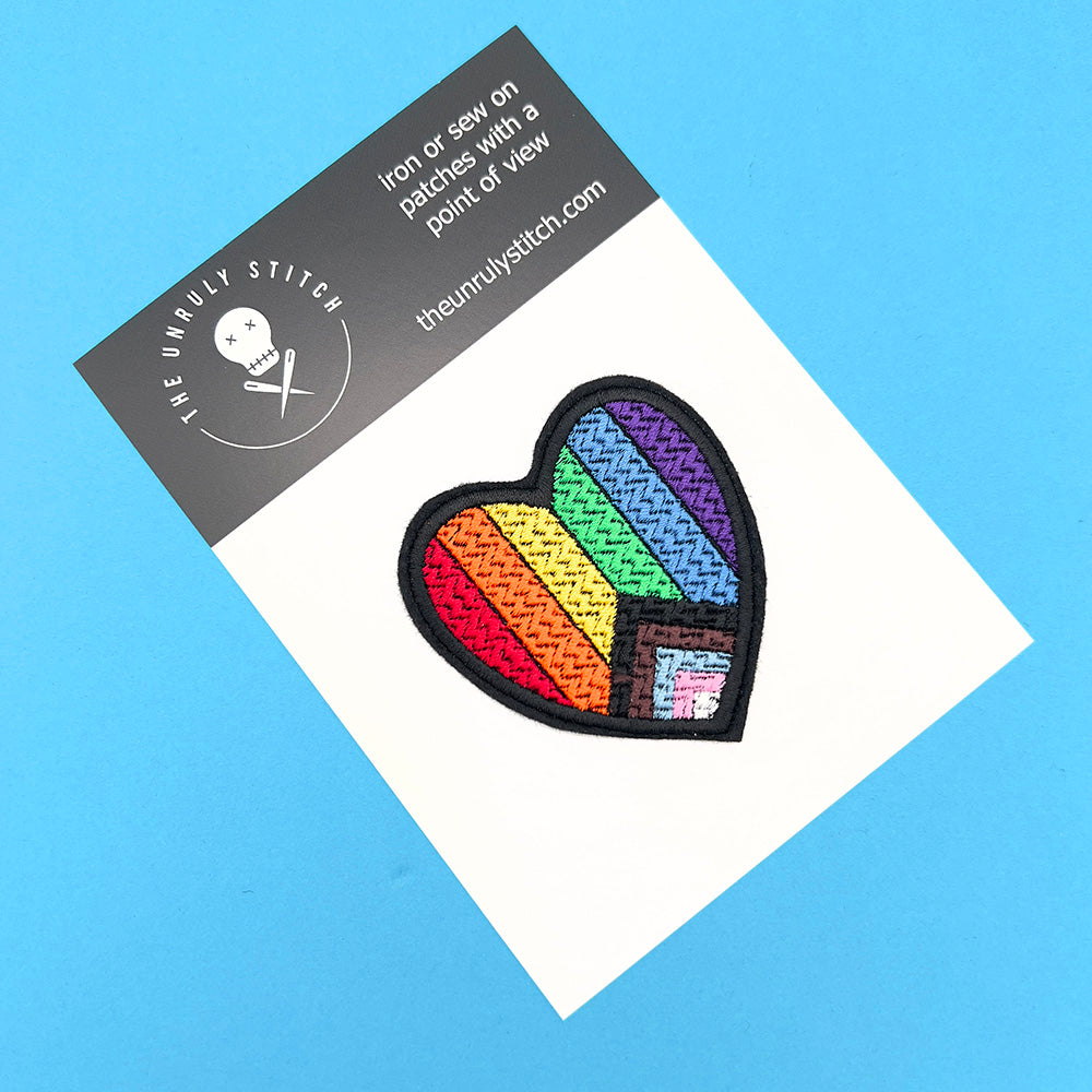 Heart-shaped embroidered inclusive Pride flag patch displayed on a branded card from "The Unruly Stitch," set against a blue background.