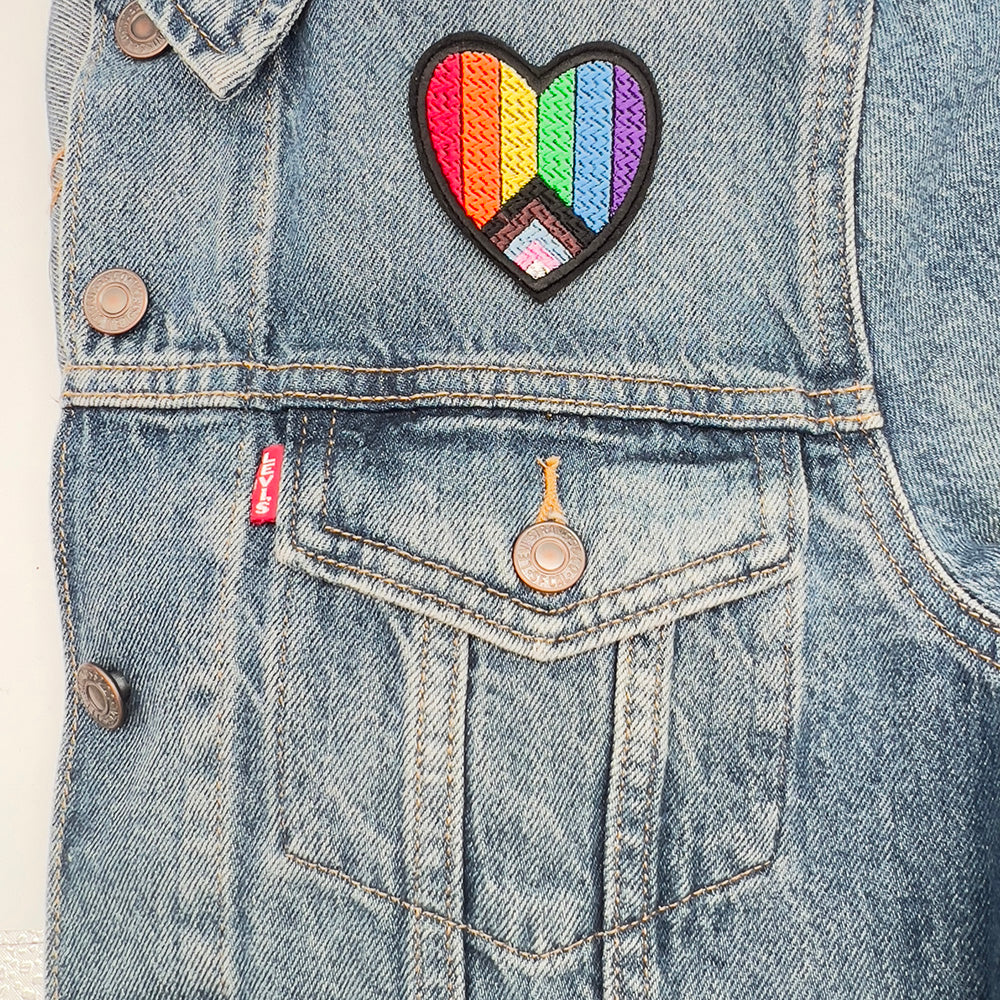 Heart-shaped embroidered inclusive Pride flag patch ironed onto the pocket of a denim jacket. The patch features horizontal rainbow stripes with an additional chevron pattern in black, brown, pink, blue, and white.