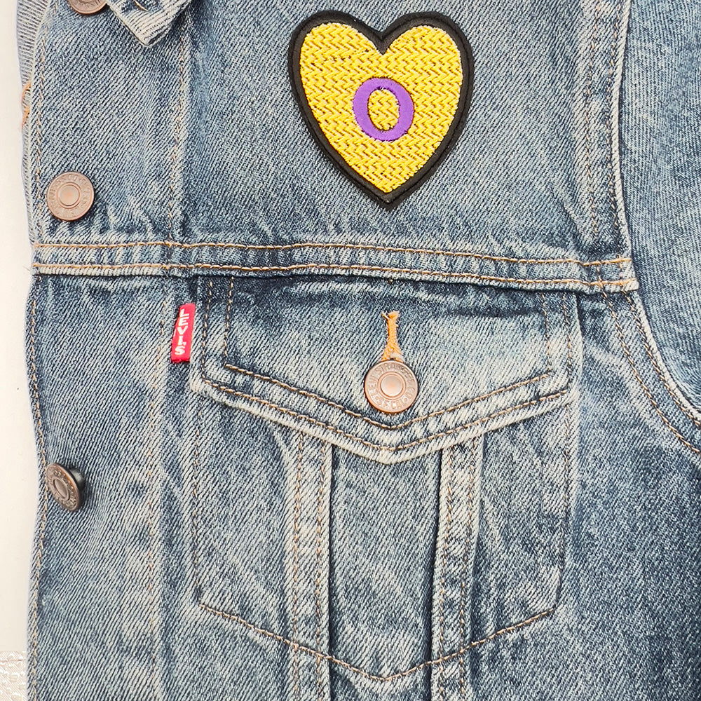 Heart-shaped Intersex Pride flag embroidered patch attached to the pocket of a denim jacket.