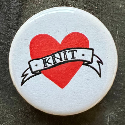 Close-up of a white pin badge with a red heart and the word "KNIT" in a banner across it