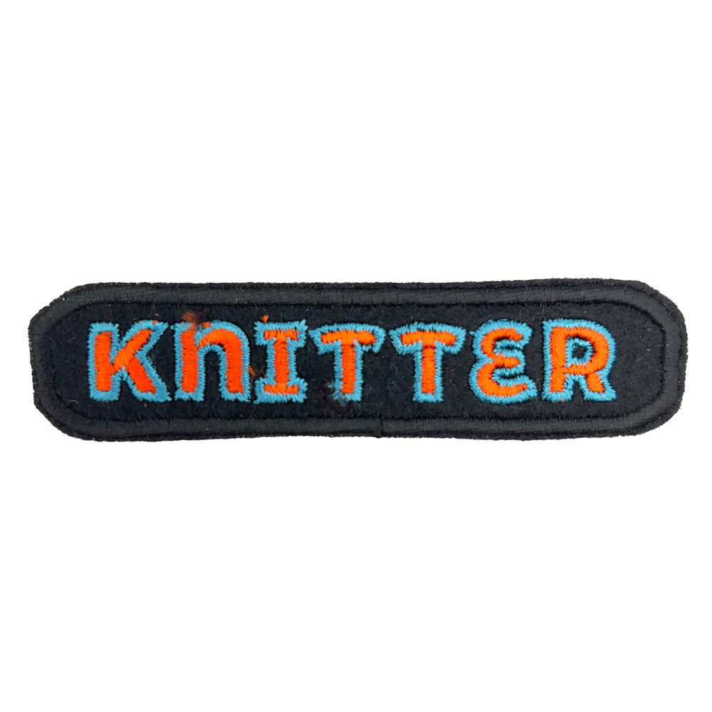 Close-up view of an embroidered felt patch with the word "Knitter." The patch features the word "Knitter" in orange letters outlined in light blue on a black background with a black border.