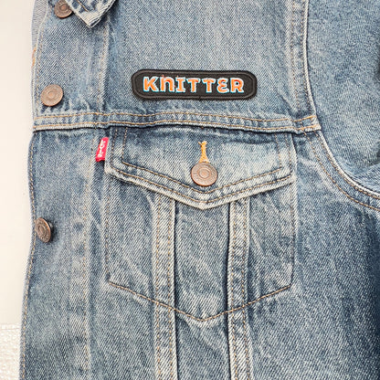 Denim jacket with an embroidered felt patch that reads "Knitter" attached above the pocket. The patch features the word "Knitter" in orange letters outlined in light blue on a black background with a black border.
