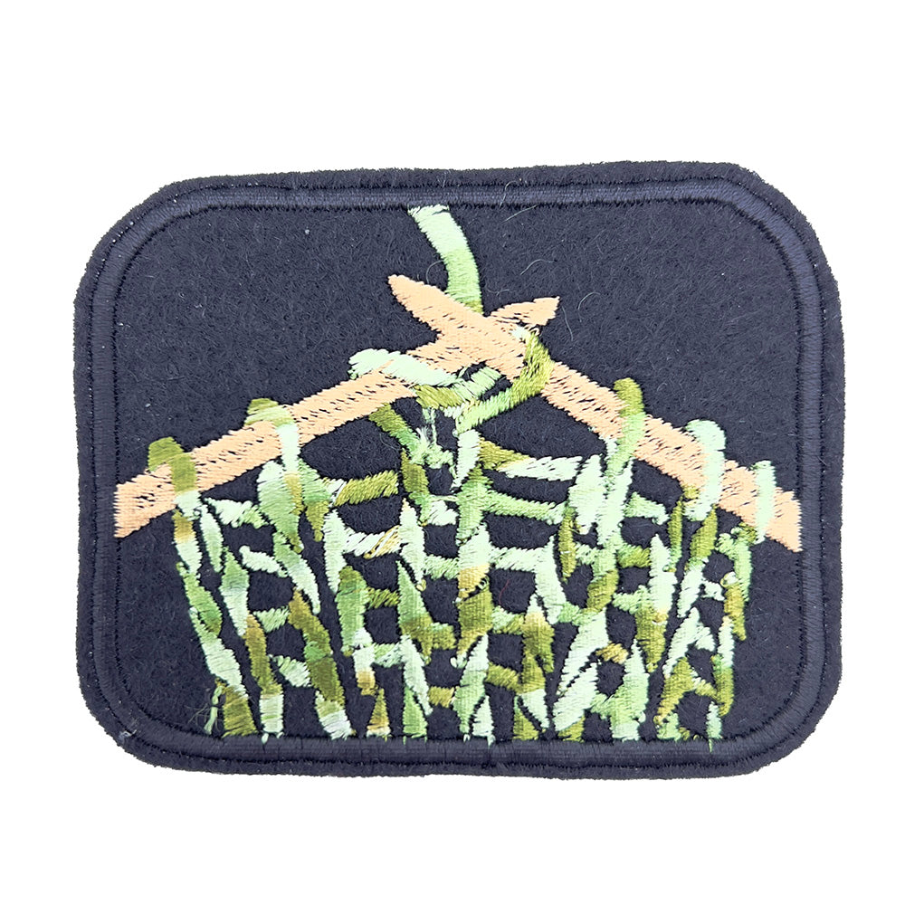 Close-up image: Close-up view of an embroidered felt patch depicting knitting on knitting needles. The patch features beige knitting needles and green, beige, and white yarn in a stocking stitch pattern against a black background.