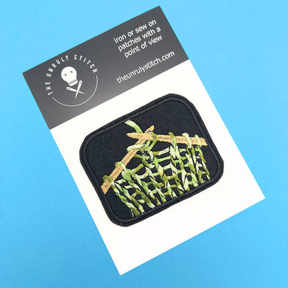 Product card image: Embroidered felt patch of knitting on knitting needles displayed on a product card. The patch features beige knitting needles and green, beige, and white yarn in a stocking stitch pattern against a black background, set on a white card with branding at the top.