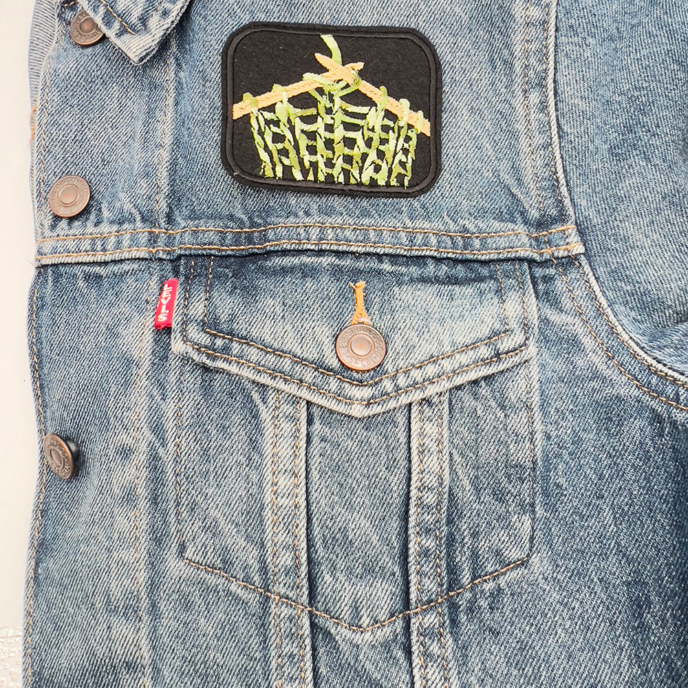 Denim jacket image: Embroidered felt patch of knitting on knitting needles affixed to a denim jacket. The patch features beige knitting needles and green, beige, and white yarn in a stocking stitch pattern against a black background, positioned above the left pocket of the jacket.