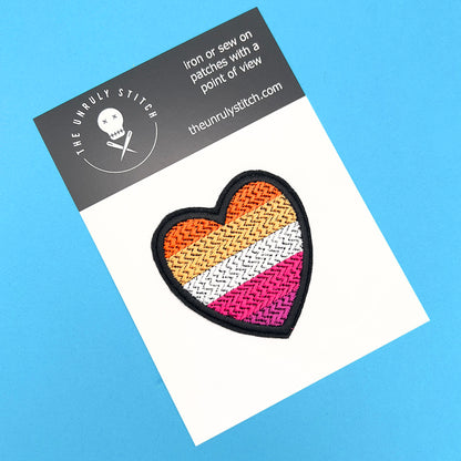 A heart-shaped embroidered felt patch with the lesbian pride flag colors attached to a card. The card has a black header with the brand's logo, "The Unruly Stitch," and the website address.