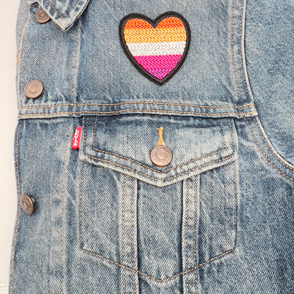 A heart-shaped embroidered felt patch with the lesbian pride flag colors affixed to the upper left part of a denim jacket.