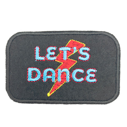 A close-up image of a rectangular embroidered felt patch with the text "LET'S DANCE" in blue and red letters, set against a black background. A red and yellow lightning bolt is embroidered behind the text.