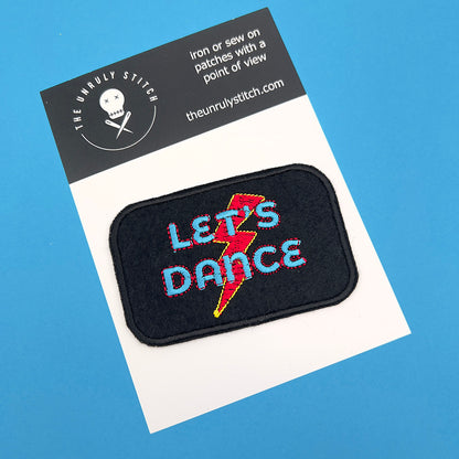 An embroidered felt patch with the text "LET'S DANCE" and a lightning bolt on a display card. The patch is ready for sewing or ironing onto fabric, with the brand "The Unruly Stitch" displayed on the card.