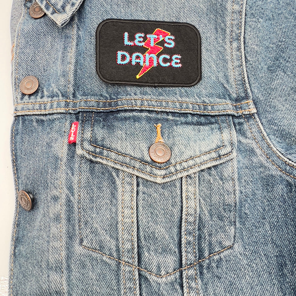 A denim jacket with a "LET'S DANCE" embroidered patch sewn on the upper pocket area. The patch features blue and red text with a red and yellow lightning bolt on a black background.
