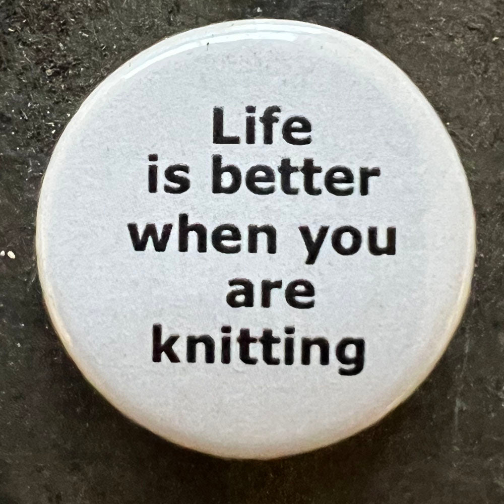 Close-up of a white Life is better when you are knitting pin badge with black text.